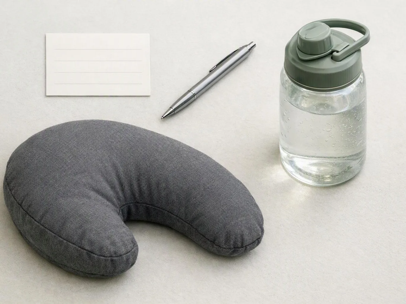 A gray travel neck pillow, silver pen, blank note card, and water bottle sit on a light tabletop