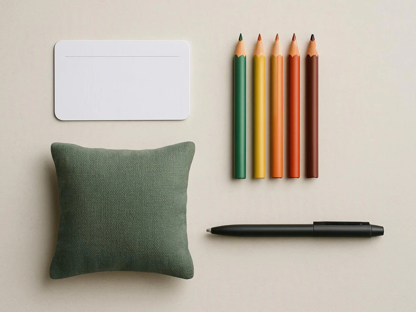 A green fabric cushion, blank card, five colored pencils, and a black pen rest on a beige surface