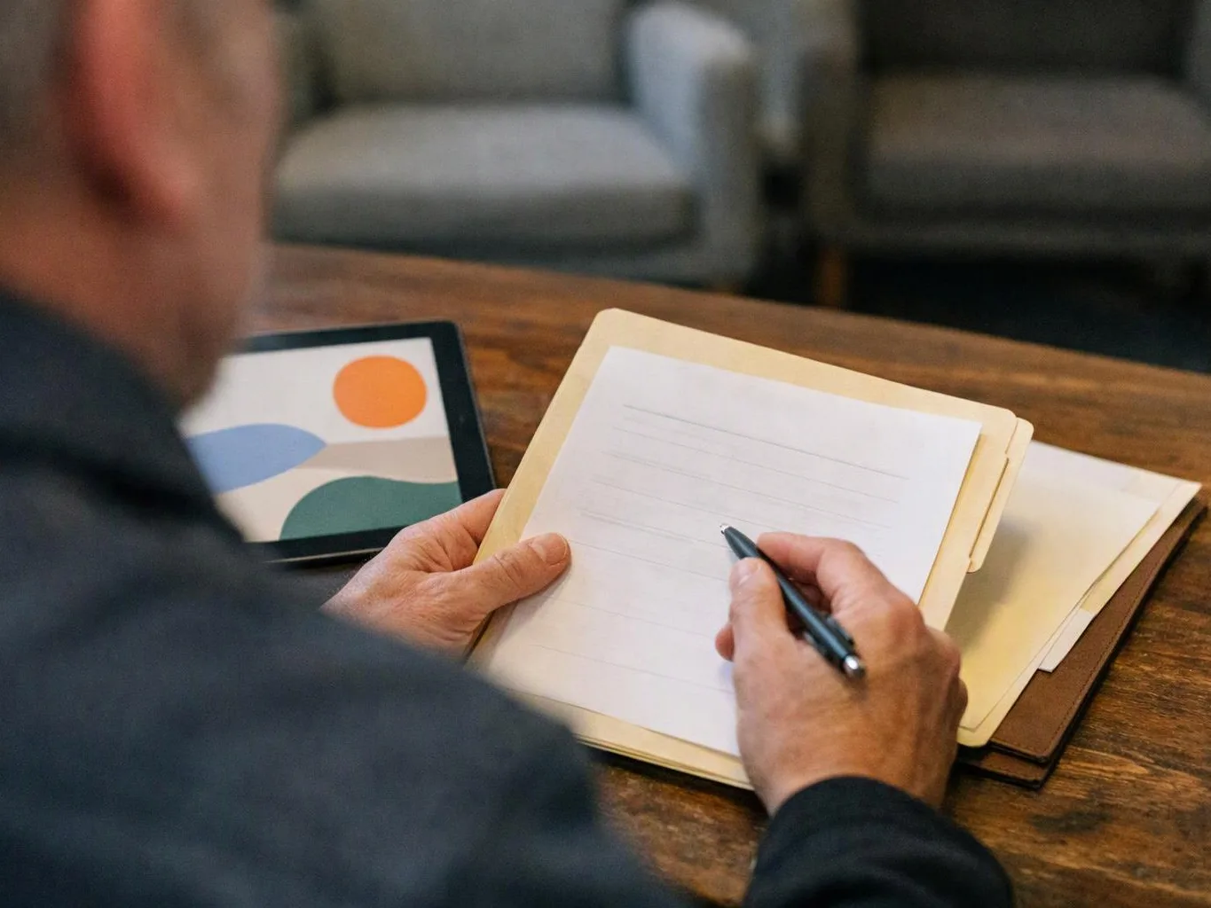 A person writes on lined paper at a wooden table beside a tablet showing simple colorful shapes